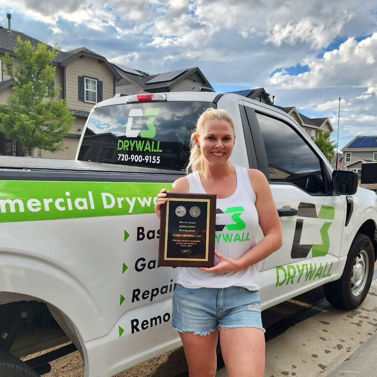 Katie Andrews with an award recognizing Core3 Drywall's sponsorship of VetFest, standing beside the C3 Drywall truck.